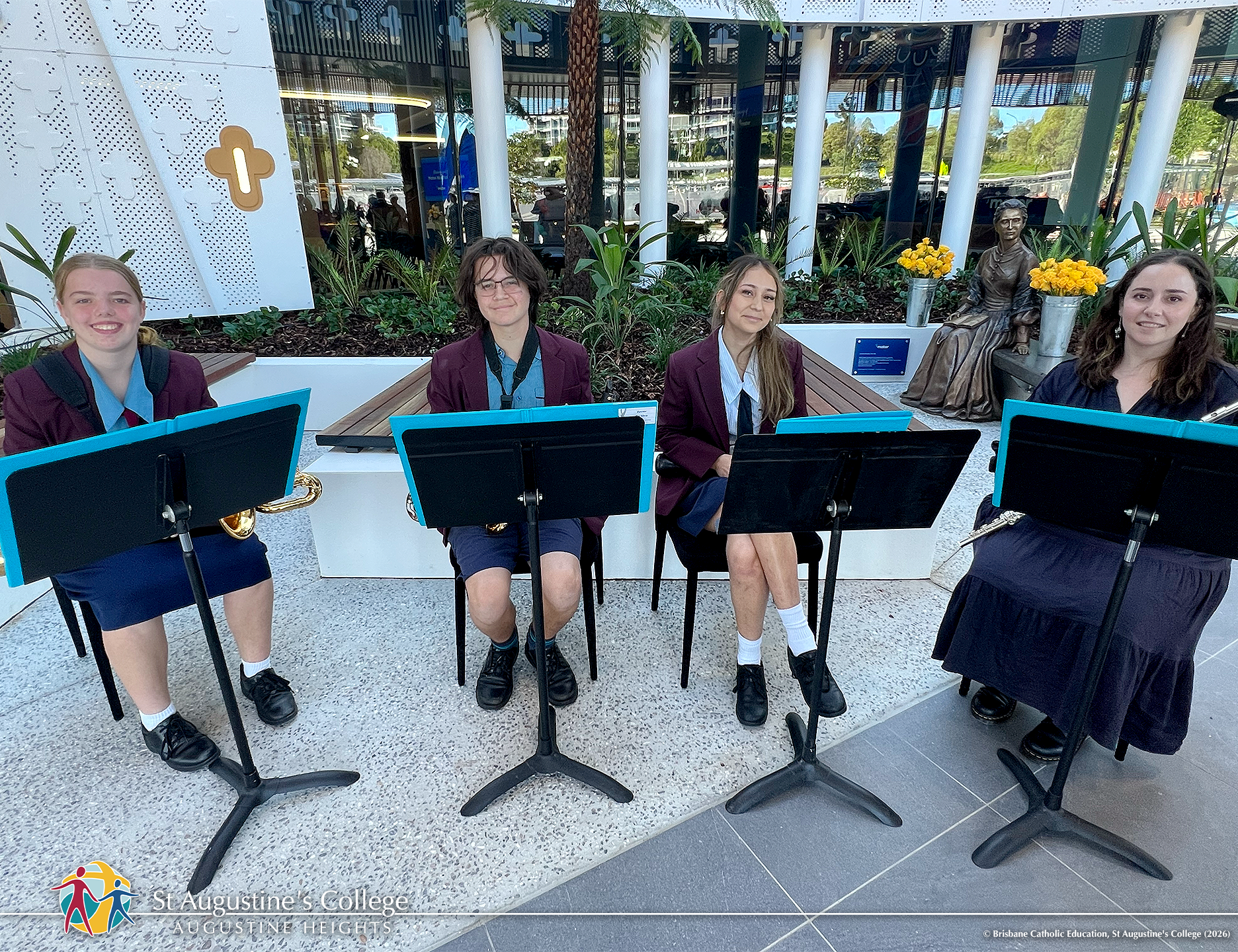 Four students in school uniform sit with music stands and instruments in an outdoor courtyard featuring plants and a patterned building façade.