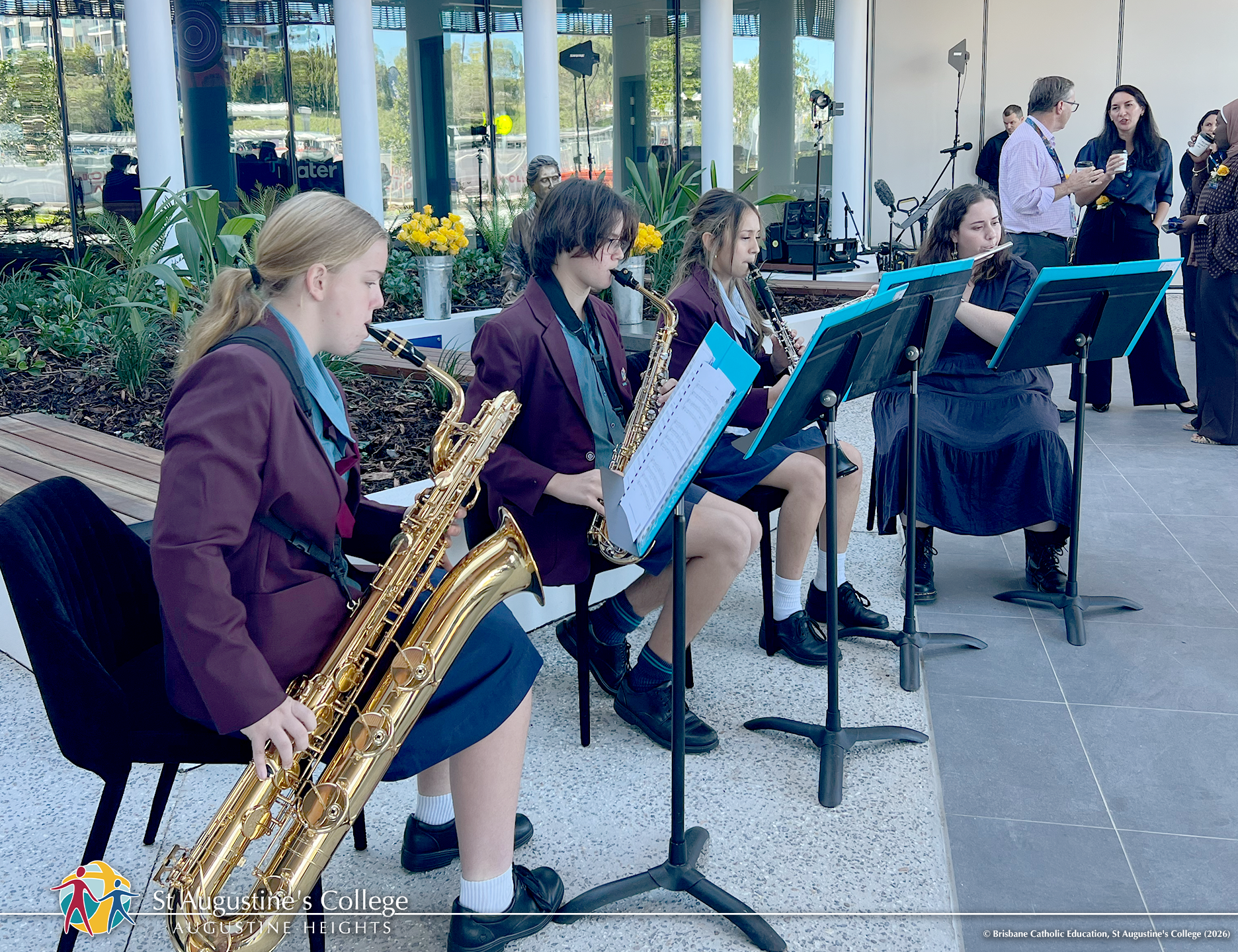 Students in school uniform play wind instruments while seated beside music stands in an outdoor setting, with staff and equipment visible in the background.