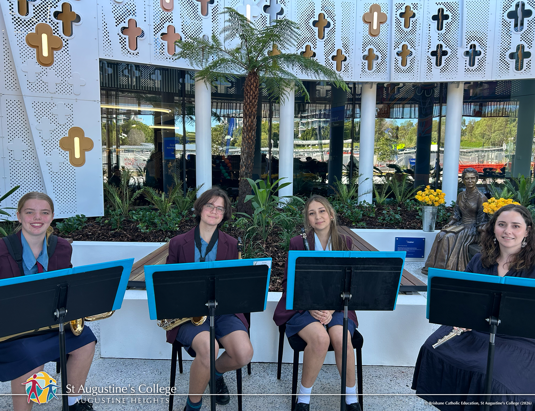 Four students in school uniform sit outdoors with music stands and instruments in front of landscaped gardens and a building with patterned panels.