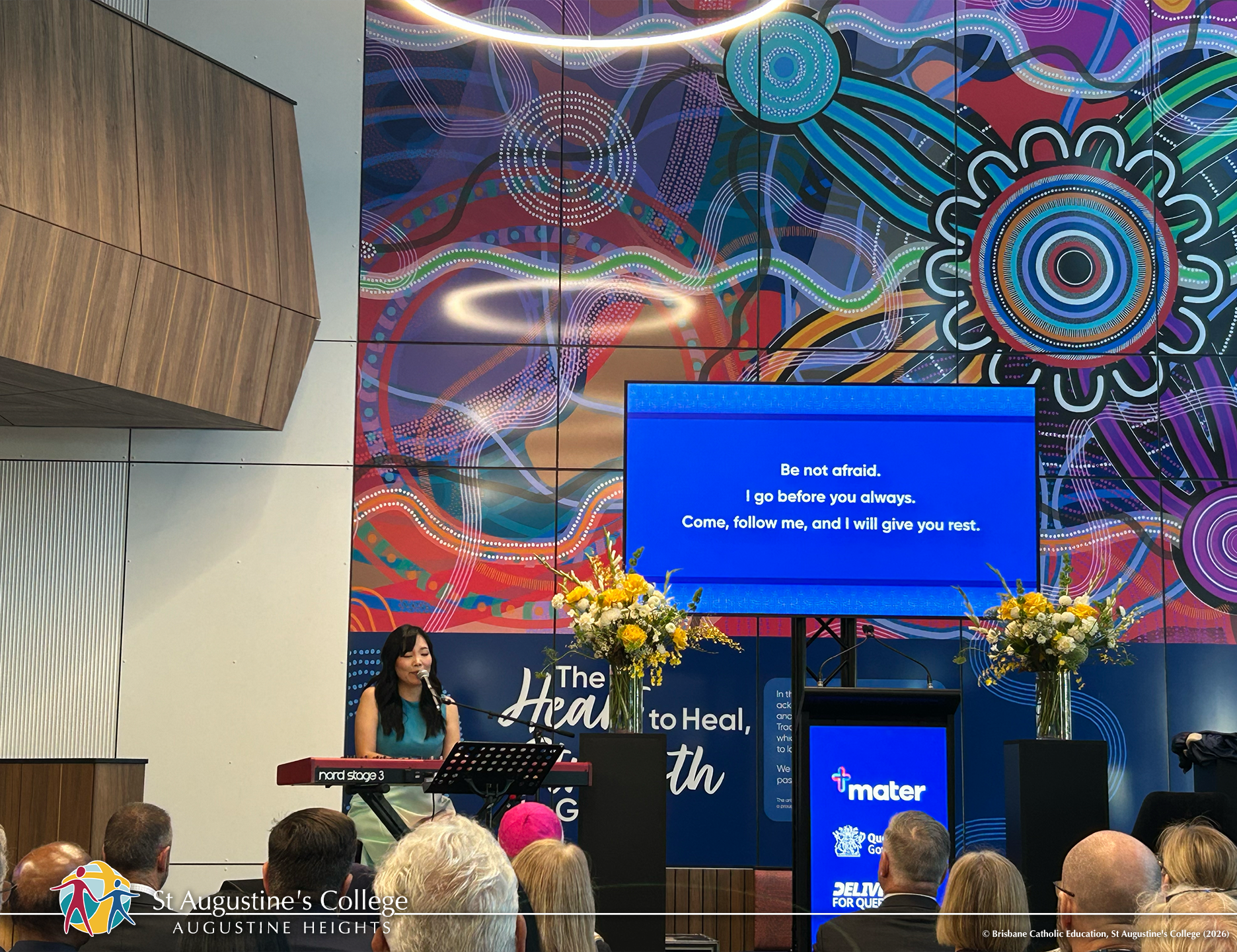 A person performs music on a keyboard at a lectern in a large indoor venue, with a screen displaying text and an audience seated in front.