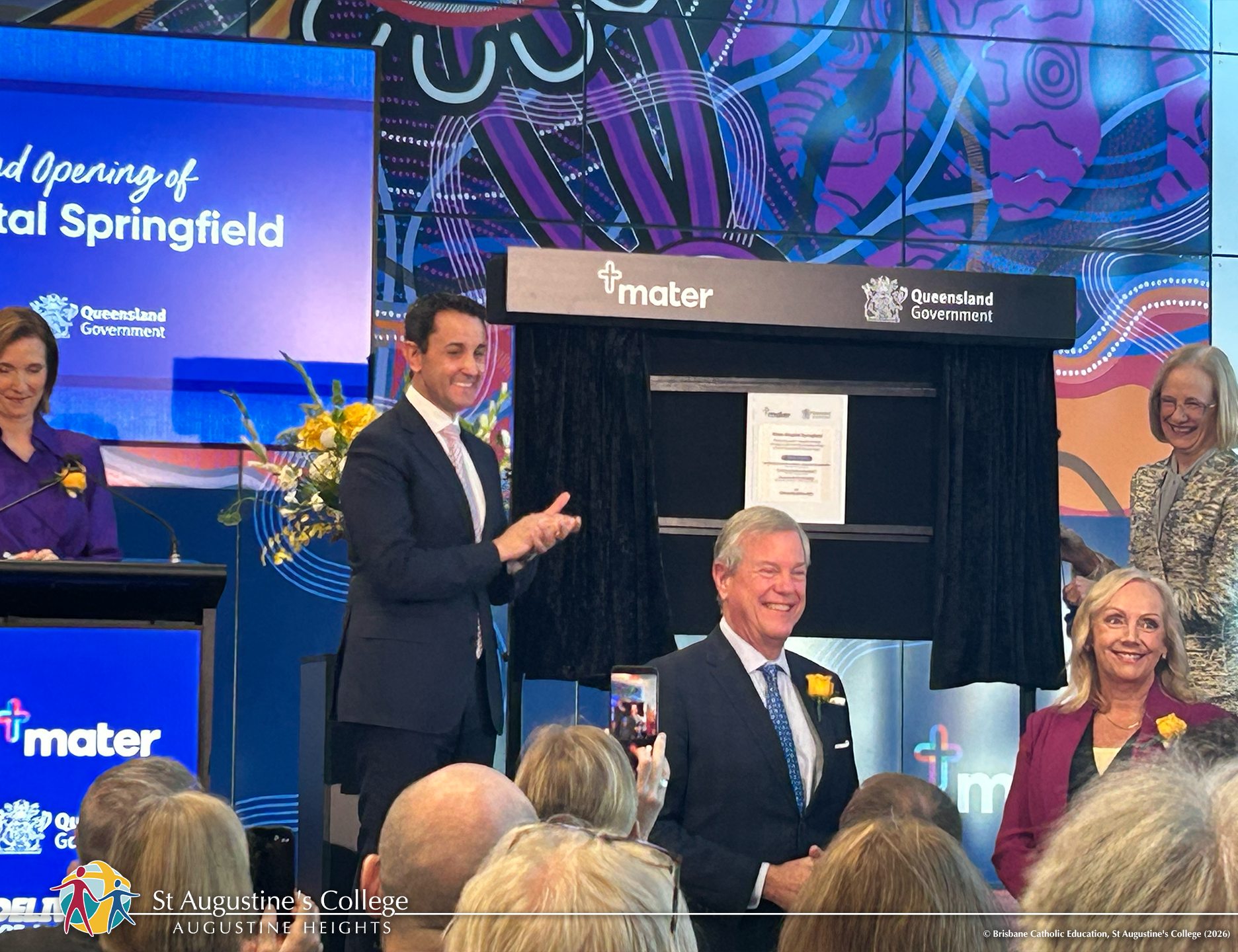 People stand on a stage during a formal event, applauding beside a covered plaque and display boards at a venue branded with Mater and Queensland Government signage.