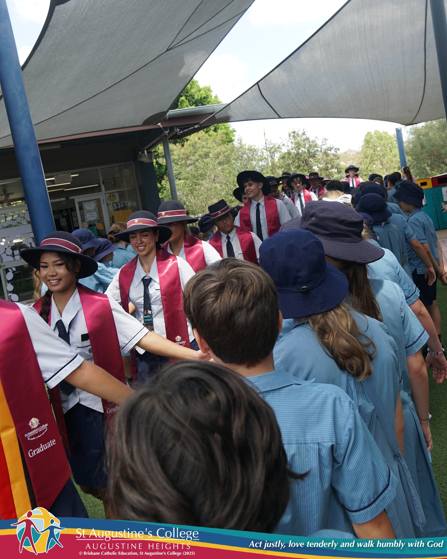 Senior students in formal uniform with red graduation stoles walk through lines of younger students in blue uniforms under shaded outdoor walkways, greeting them during a school procession.